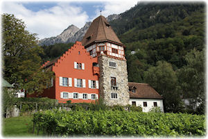 Die Architektur der Bauten weist einen ganz besonderen Stil auf - Das rote Haus. Ein turmartiges, rotes Haus samt Nebengebäuden vor einem Gebirge.